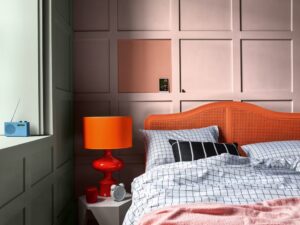 Geometric orange and peach feature wall behind a bedframe with an orange velour headboard and bold orange accent lamp. 
